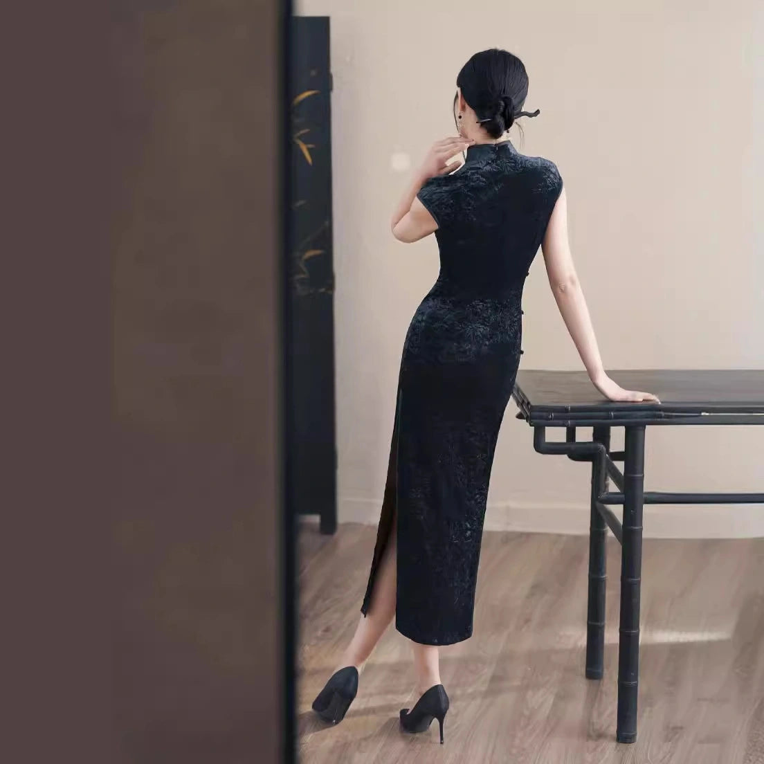 Woman in a Black Floral Velvet Qipao Cheongsam Dress  standing by a table in a room with wooden flooring.