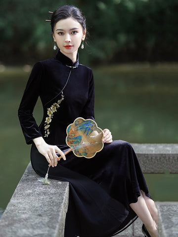 A woman sits on a stone ledge by a body of water, wearing a black long-sleeve velvet a Line cheongsam-style dress with gold floral embroidery near the shoulder. She holds a decorative fan with a blue and gold landscape design.