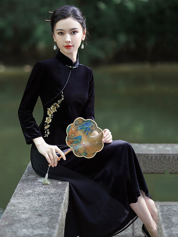 A woman sits on a stone ledge by a body of water, wearing a black long-sleeve velvet a Line cheongsam-style dress with gold floral embroidery near the shoulder. She holds a decorative fan with a blue and gold landscape design.