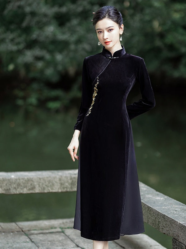 A woman wearing a long black velvet a line qipao cheongsam-style dress with gold embroidery, standing on a stone bridge with a blurred green outdoor background.