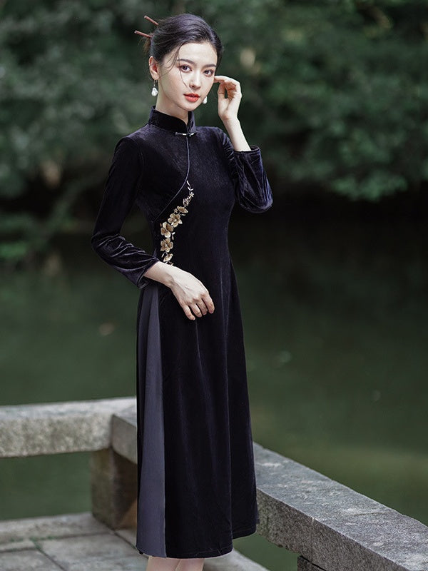 A woman wearing a black velvet long-sleeve a line qipao cheongsam dress with gold embroidery on the side, standing on a stone bridge with a blurred green garden background.
