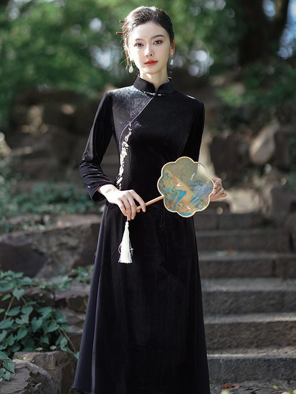 A woman wearing a black long-sleeve traditional a line qipao dress with a silver embroidered side panel and holding a decorative fan, standing on stone steps in a garden setting.
