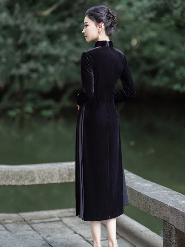 A woman wearing a long black velvet a line cheongsam dress with a high mock neck and long sleeves, standing on a stone bridge with a green pond and trees in the background.