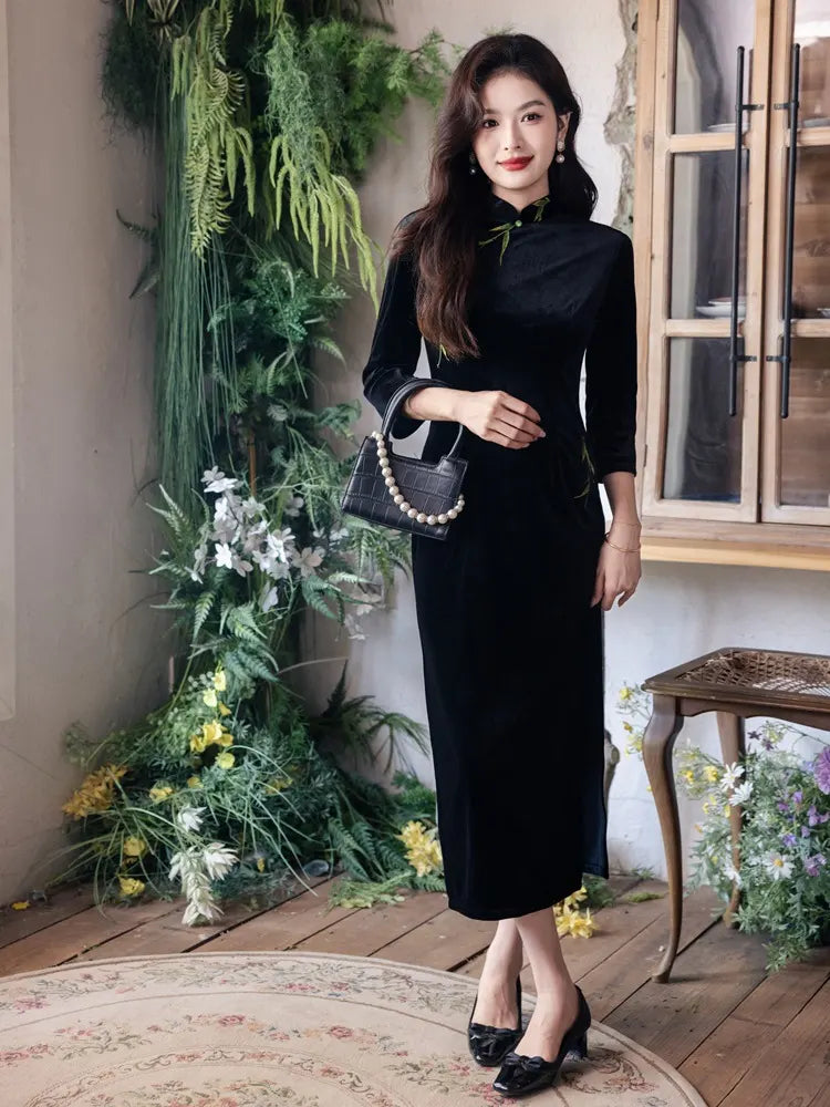 Woman in a Black Long Sleeves Qipao Dress with Green Embroidery holding a handbag in a decorated room with plants and flowers.