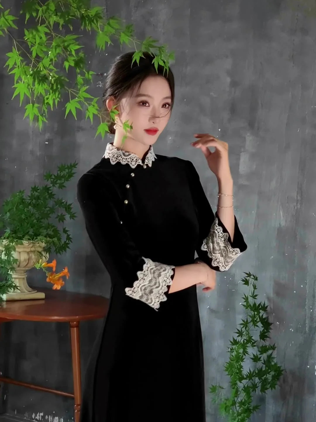 Woman in a Black Long Sleeves Velvet A-Line Qi pao with Lace Trim details against a gray background with green plants.