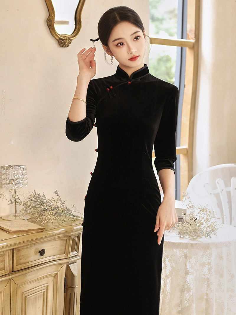 A woman wearing a black velvet cheongsam-style long dress with red button accents, standing in a softly lit indoor room with a cream wall, mirror, cabinet, and table visible.