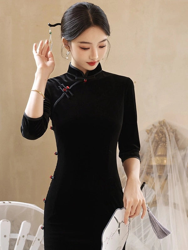 A woman wearing a black velvet cheongsam-style dress with red decorative accents, holding a white folding fan in a softly lit indoor setting.