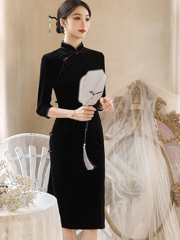 A woman models a black velvet cheongsam-style dress with three-quarter sleeves and red decorative accents on the collar and front. She holds a white folding fan in an elegant indoor setting with a white chair and a draped veil in the background.