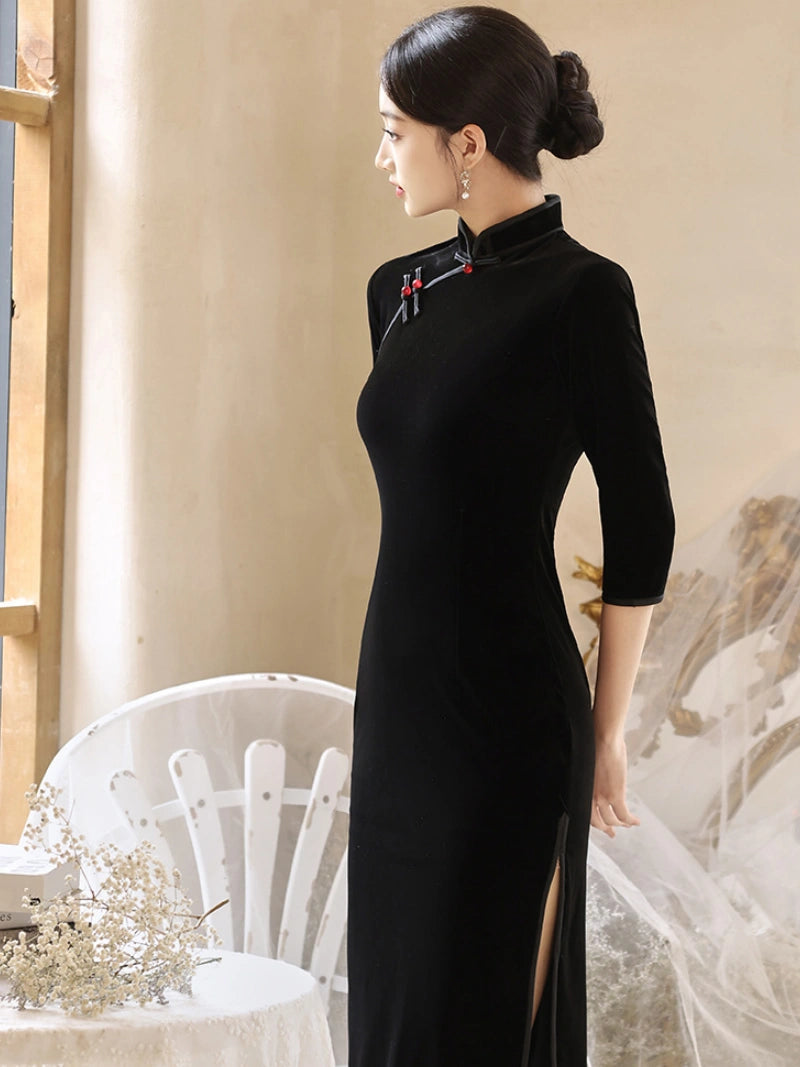 A woman wearing a black velvet cheongsam dress with a high collar, three-quarter sleeves, and a side slit, standing in a softly lit indoor setting.