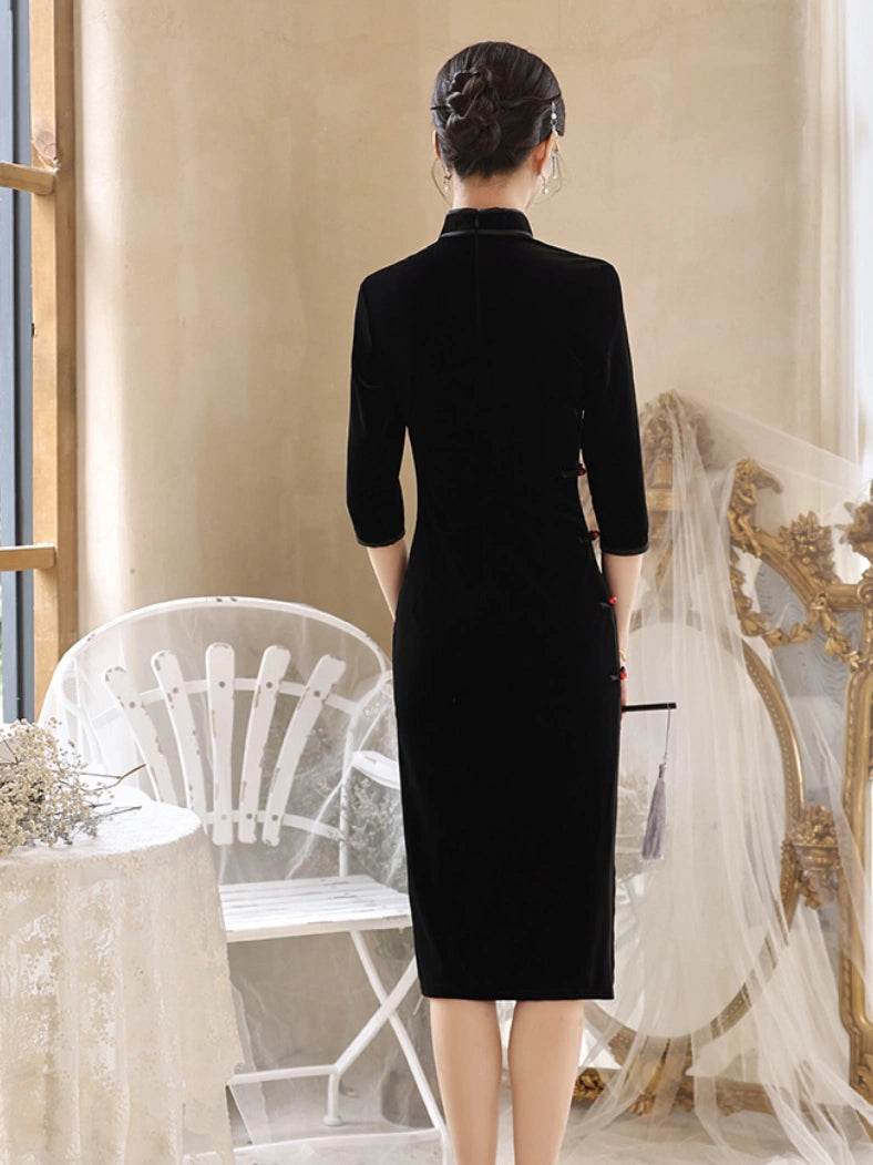A woman stands with her back to the camera wearing a black velvet midi dress with three-quarter sleeves and a high collar, styled in a vintage-inspired bridal room setting with white lace chairs and a gold mirror.