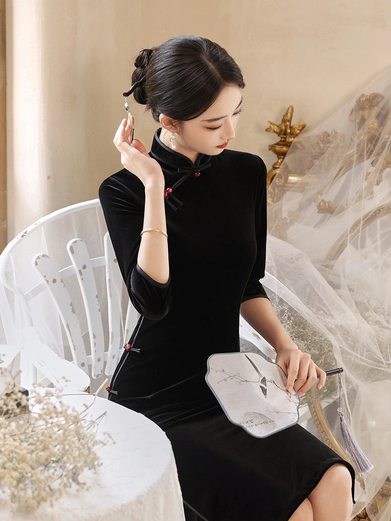 A woman wearing a black velvet cheongsam-style dress with red button accents, seated in a white chair and holding a folding fan in a softly decorated indoor setting.