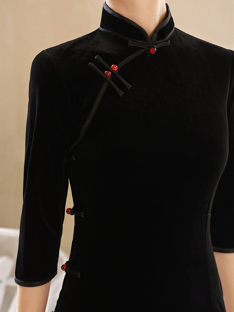 A close-up of a black velvet cheongsam-style dress or tunic with a high collar, diagonal front closure, and red decorative buttons, shown on a mannequin against a soft beige background.