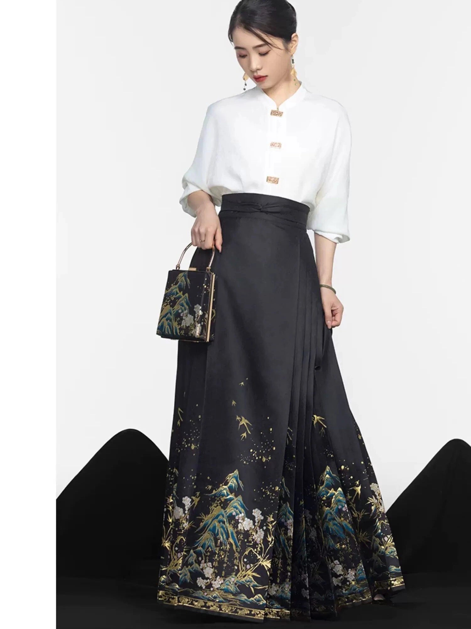A woman models a traditional Chinese-inspired outfit featuring a white mock-neck blouse with gold buttons and a long black satin pleated mamian horse face skirt with gold, blue, and white floral mountain patterns. She is holding a matching handbag against a white studio background.