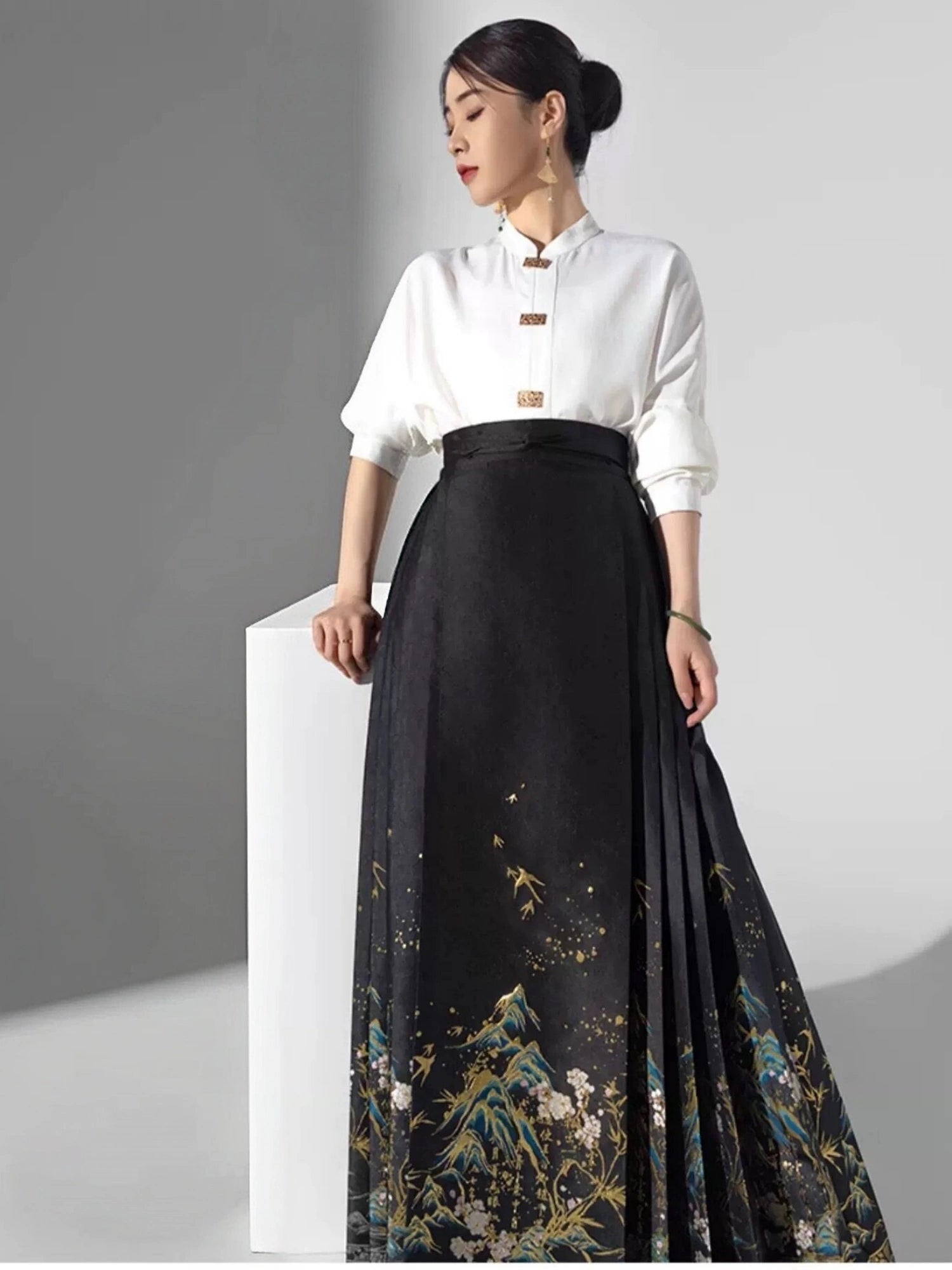 A woman models a traditional-inspired outfit featuring a white mock-neck blouse with gold buttons and a long black pleated chinese ming dynasty mamian horse skirt with a gold and blue mountain pattern, standing next to a white pedestal.