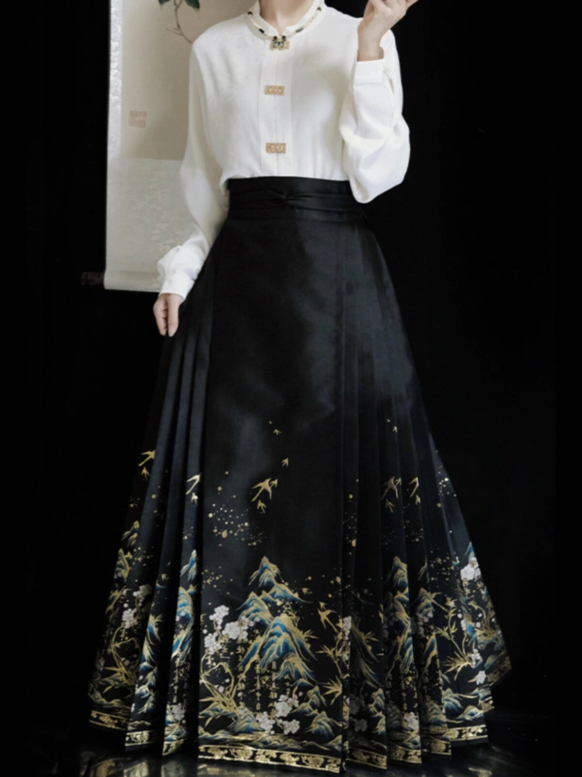 A model wearing a traditional Chinese Hanfu outfit featuring a white long-sleeve blouse with gold buttons and a high-waisted black pleated ming dynasty mamian horse face  skirt with gold, blue, and white landscape embroidery.