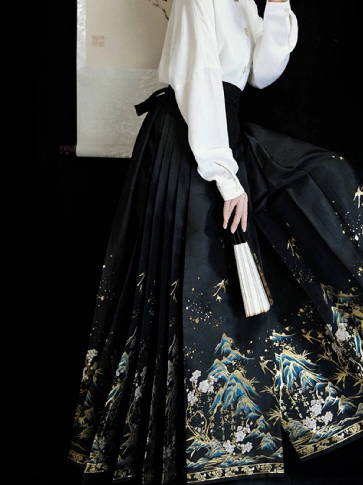 A model wearing a traditional black Hanfu mamian horse face skirt with intricate gold, blue, and white landscape embroidery, paired with a white long-sleeve blouse, holding a folding fan against a dark background.