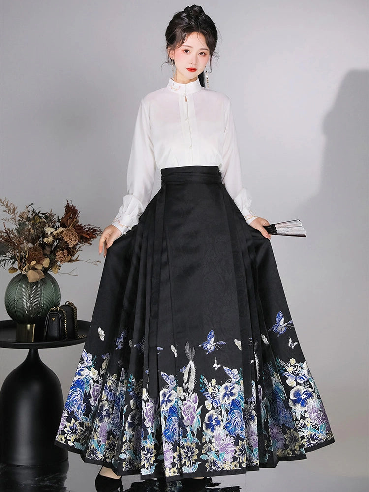 A woman models a traditional Chinese Hanfu outfit consisting of a white long-sleeve mock-neck blouse and a high-waisted black pleated mamian horse face skirt with blue and purple floral embroidery near the hem, set against a gray studio background.