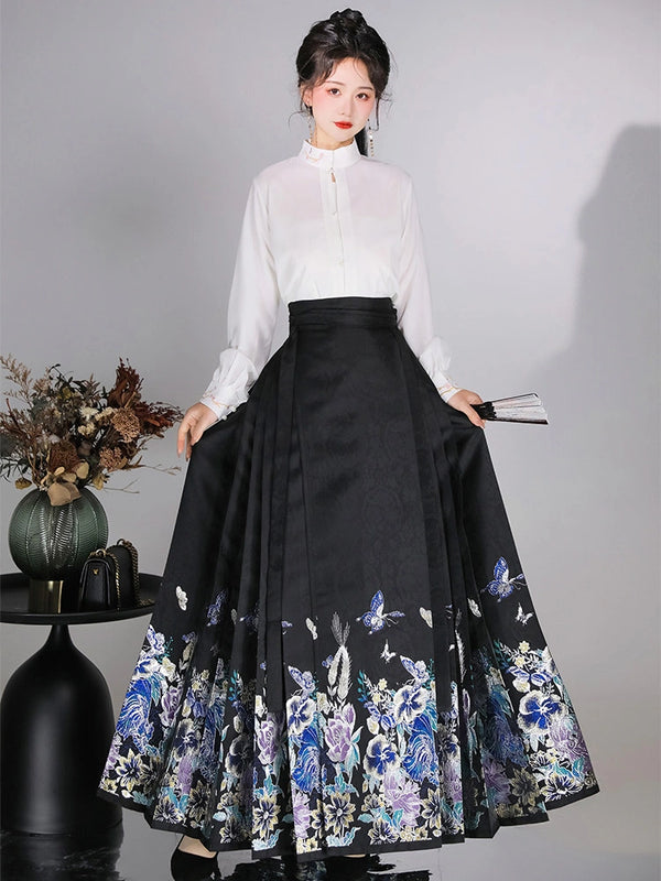 A woman models a traditional Chinese Hanfu outfit consisting of a white long-sleeve mock-neck blouse and a high-waisted black pleated mamian horse face skirt with blue and purple floral embroidery near the hem, set against a gray studio background.