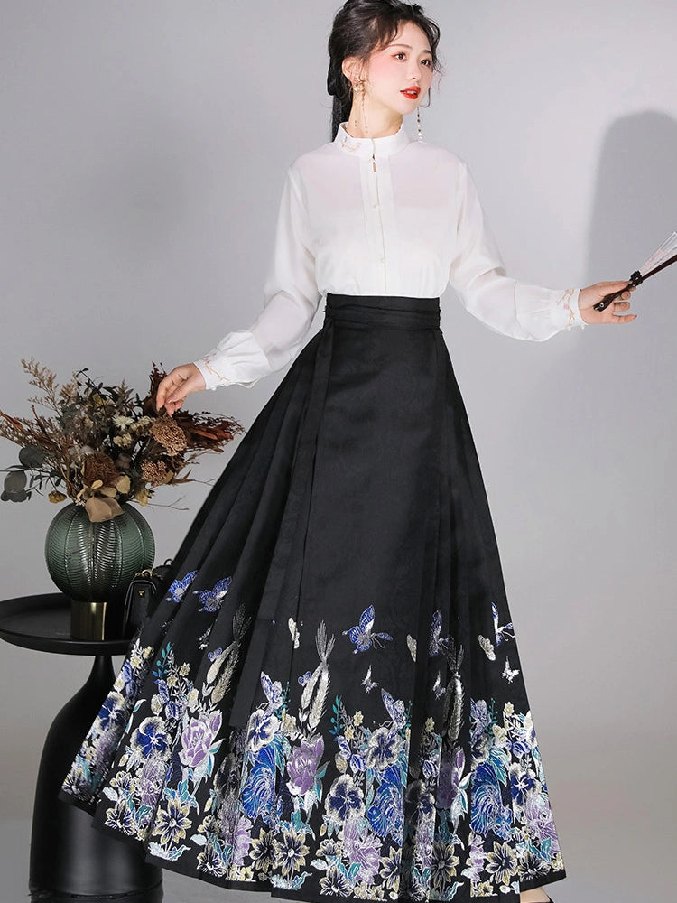 A woman models a traditional Chinese Hanfu outfit consisting of a white long-sleeve blouse and a black pleated mamian horse face skirt with blue and purple floral embroidery and butterfly motifs near the hem. She is standing against a light gray studio background next to a small black table with a decorative vase of dried flowers.
