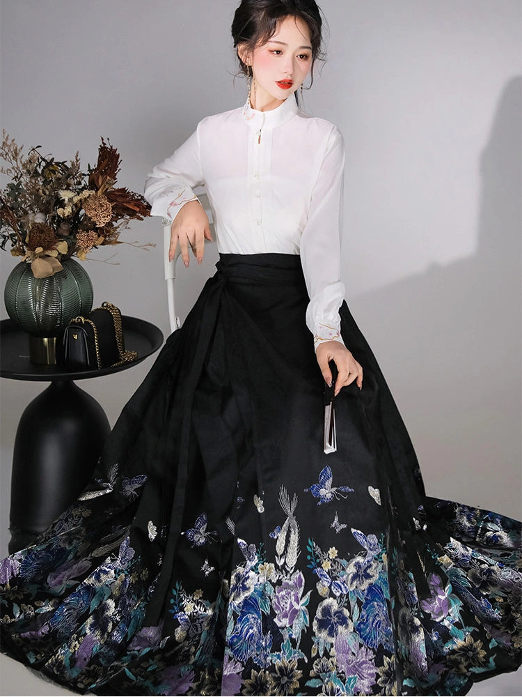 A woman models a high-neck white long-sleeve blouse paired with a voluminous black mamian horse face  skirt featuring a blue and purple floral and butterfly print, posed next to a decorative side table.