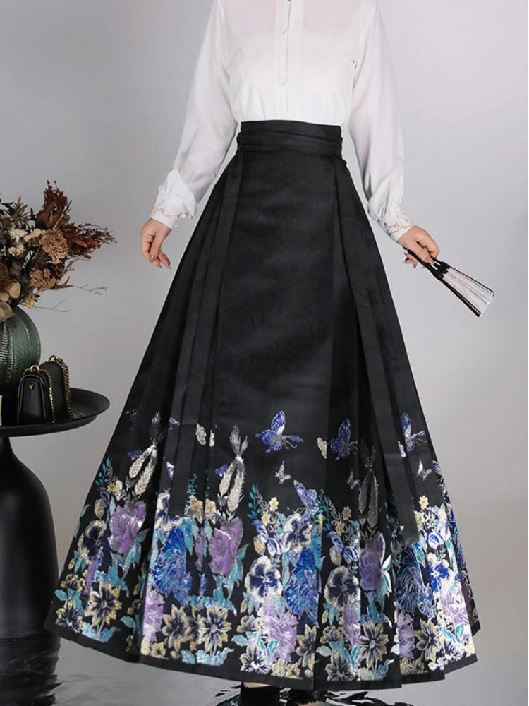 A model wearing a high-waisted black pleated mamian horse face skirt with a vibrant blue, purple, and gold floral and butterfly pattern at the hem, paired with a white blouse.