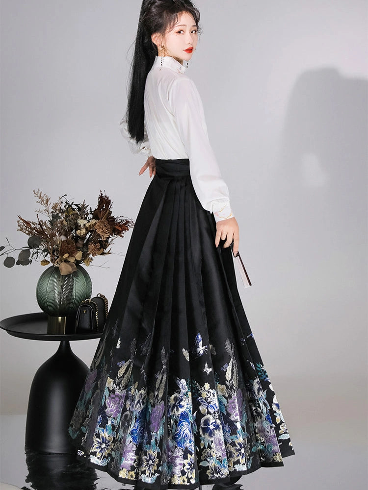 A woman models a long black mamian horse face skirt with a colorful floral and butterfly pattern along the hem, paired with a white long-sleeve blouse, in a studio setting with decorative dried flowers on a side table.