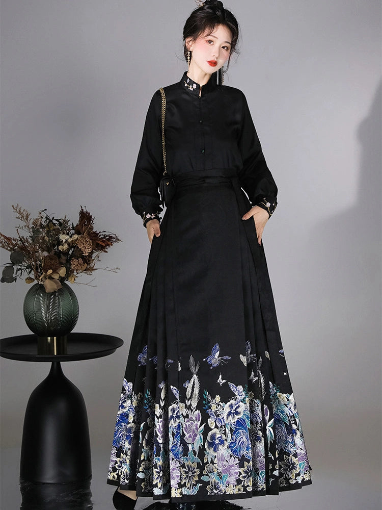 A woman models a long black dress with a high collar, button front, and a wide skirt featuring a blue and white floral print at the hem. She is standing against a gray studio background next to a small black side table with a vase of dried flowers.