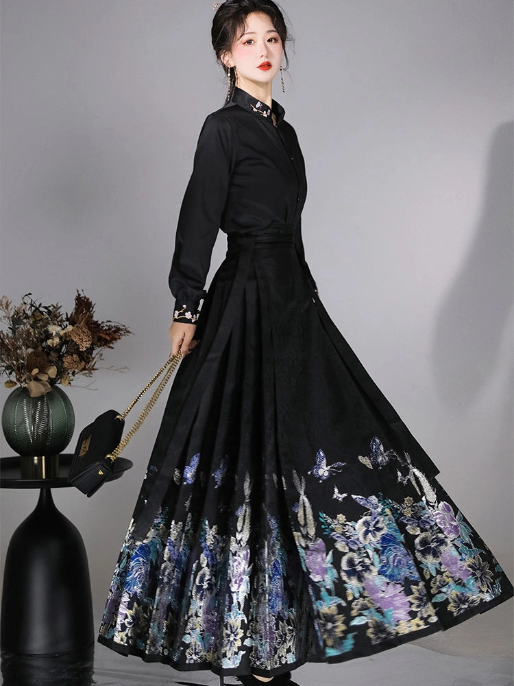 A woman models a long-sleeved black dress with a high collar and a dramatic, wide flared mamian horse face skirt featuring a colorful floral and butterfly pattern at the hem. She is holding a black handbag with a gold chain strap against a neutral gray studio background.
