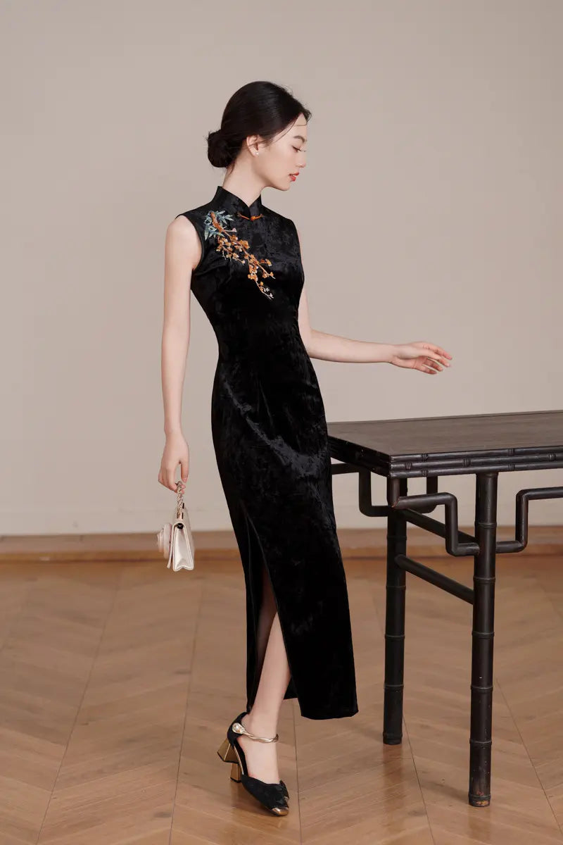 Woman in aBlack Modern Velvet Sleeveless Embroidered Qipao  with floral embroidery standing next to a wooden table.