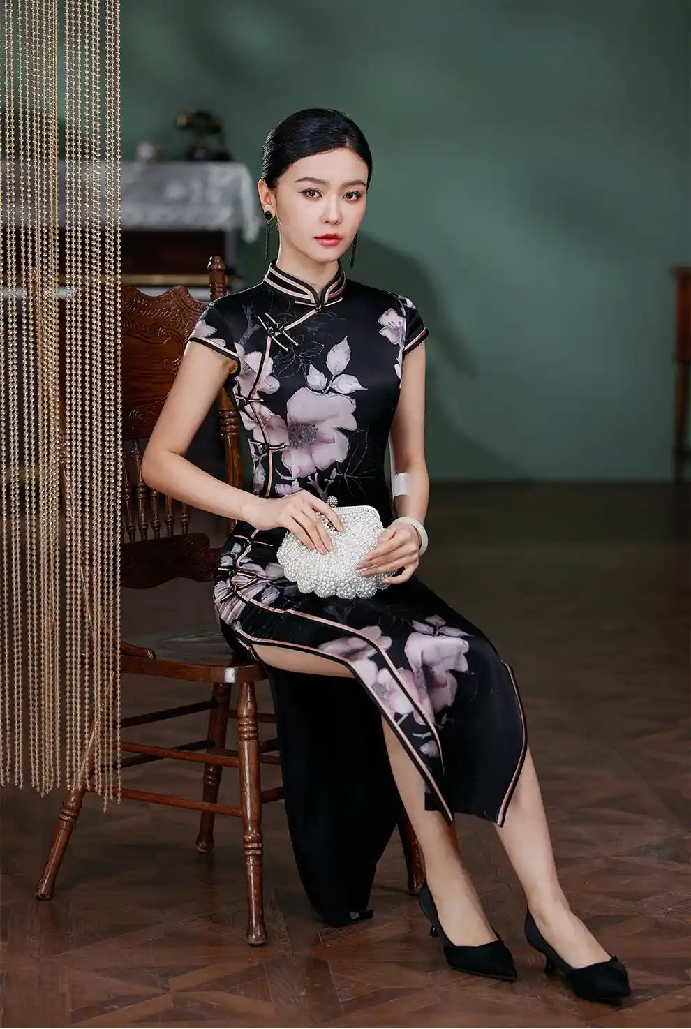 Woman in a Black Silk Chinese Qipao Dress – Elegant Mandarin Collar Cheongsam  sitting on a wooden chair in a room with a green wall.