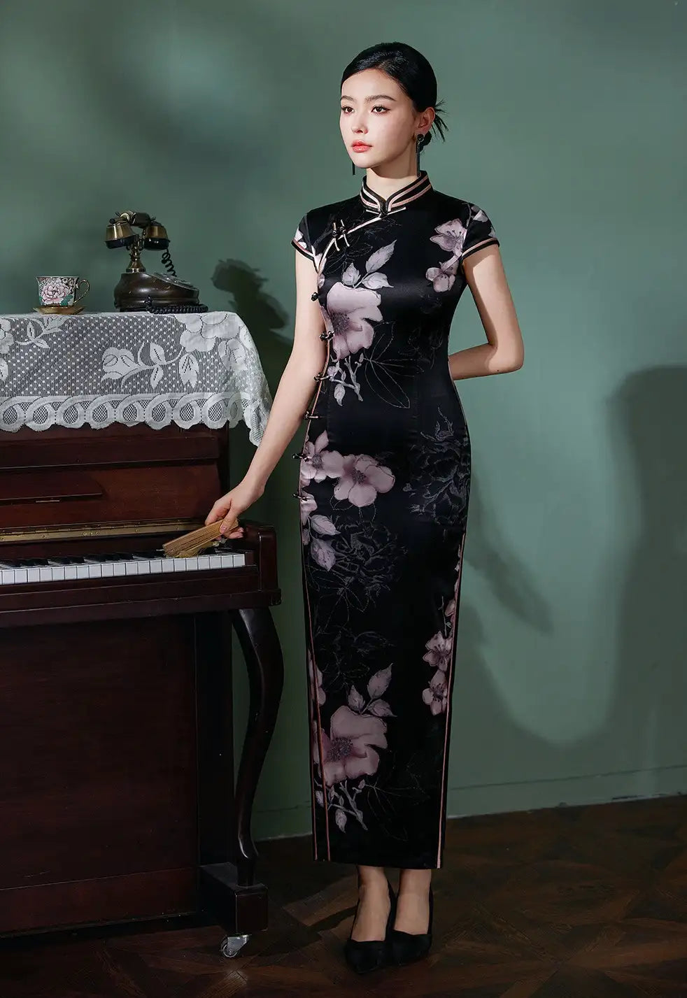 Woman in a Black Silk Chinese Qipao Dress – Elegant Mandarin Collar Cheongsam standing next to a piano against a green wall.