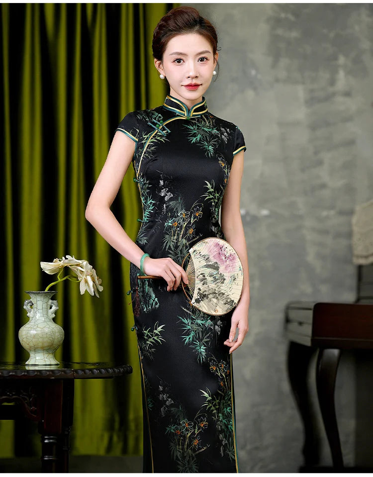Woman in a black floral  Silk Qipao cheongsam dress with Bamboo & Orchid Print  holding a fan against a gray and green curtain background