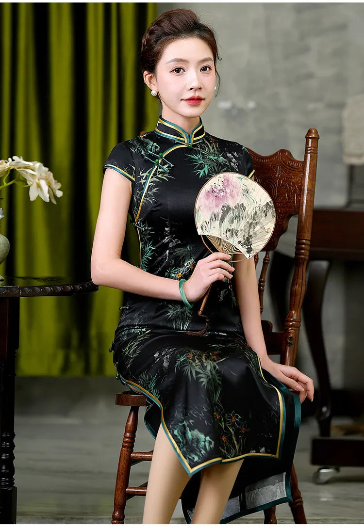 Woman in a traditional black and green Silk Qi pao with Bamboo & Orchid Print  holding a fan, sitting on a wooden chair.