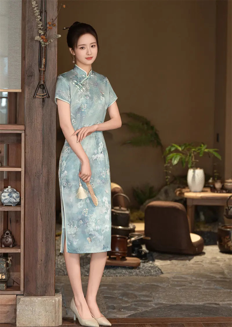 Woman in a light Blue Floral Cheongsam Qipao Dress standing in a room with wooden furniture and plants.