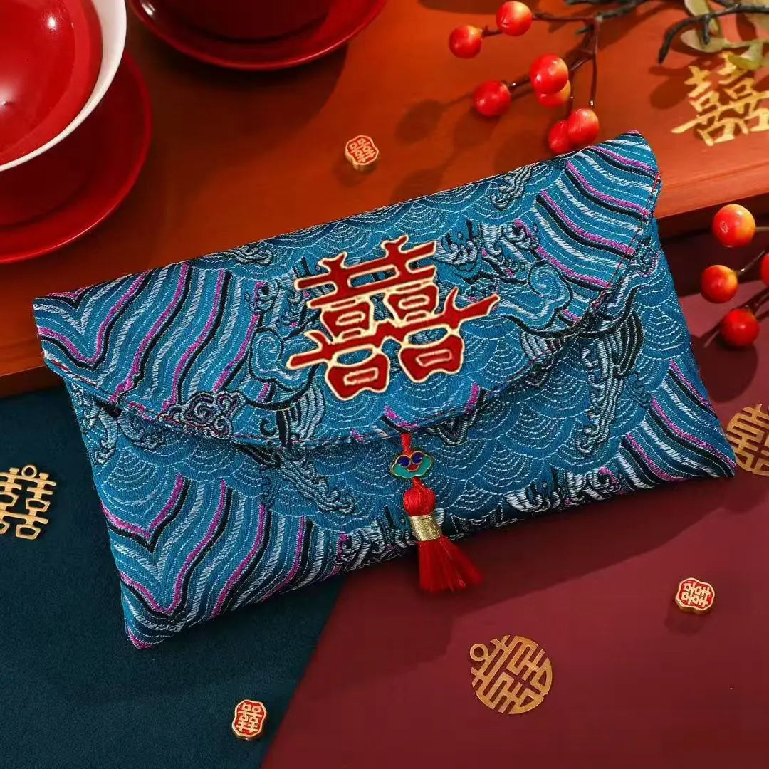 A blue embroidered envelope-style Chinese red packet (hongbao) with a red double happiness symbol and tassel, styled on a wooden surface with red cups, decorative gold symbols, and red berries.