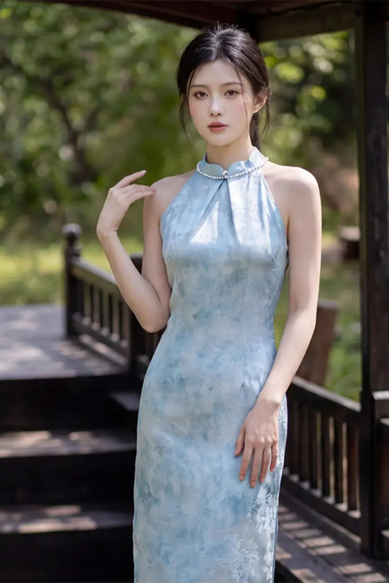 A woman wearing a light blue and white watercolor-patterned high-neck Modern Pearl-Embellished Halter-Style Qipao  cheongsam dress, posing outdoors on a wooden platform with a blurred green garden background.