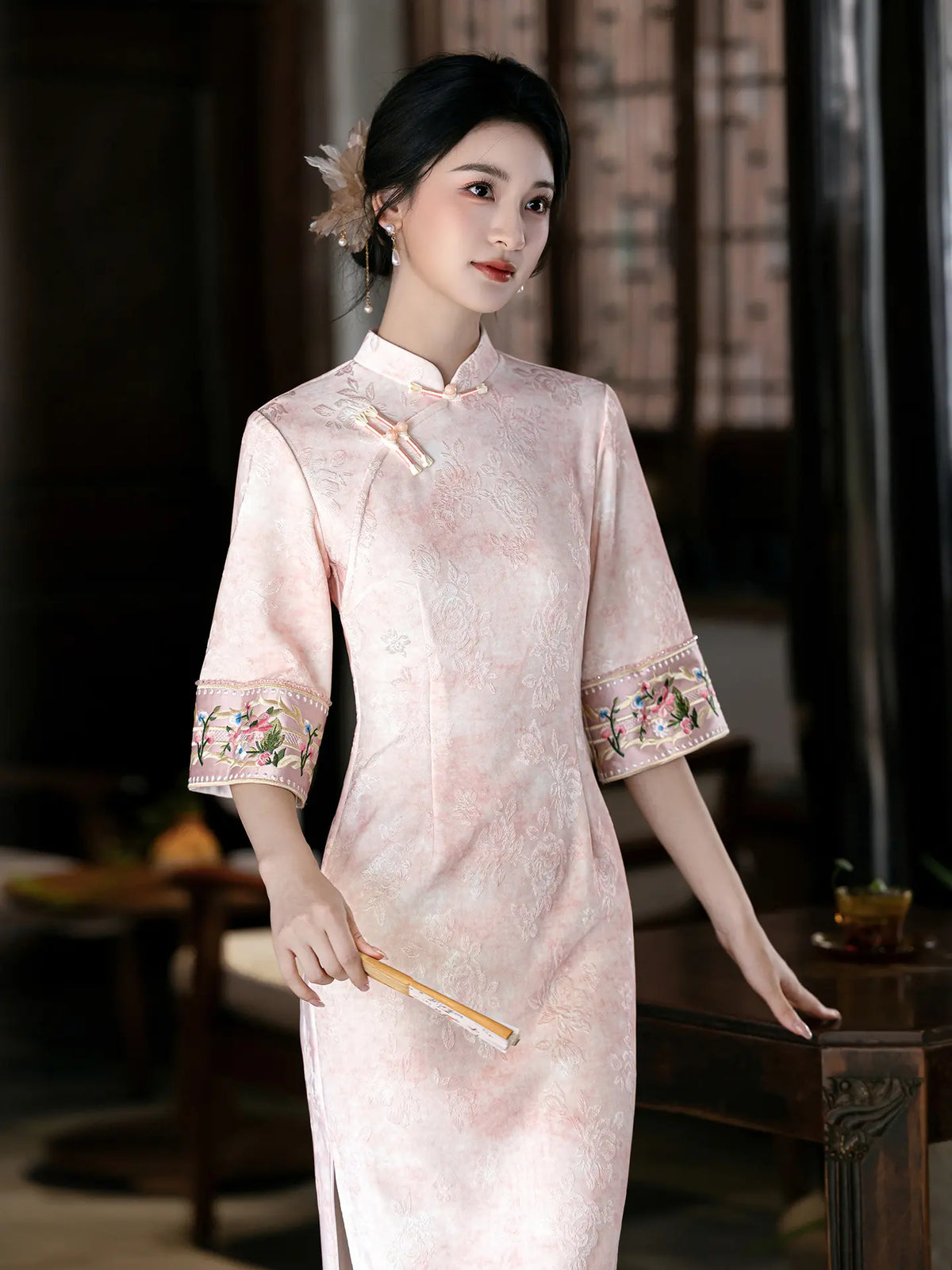 A woman wearing a pale pink traditional Chinese cheongsam dress with floral embroidery on the sleeves, standing in a dimly lit indoor setting and holding a folding fan.