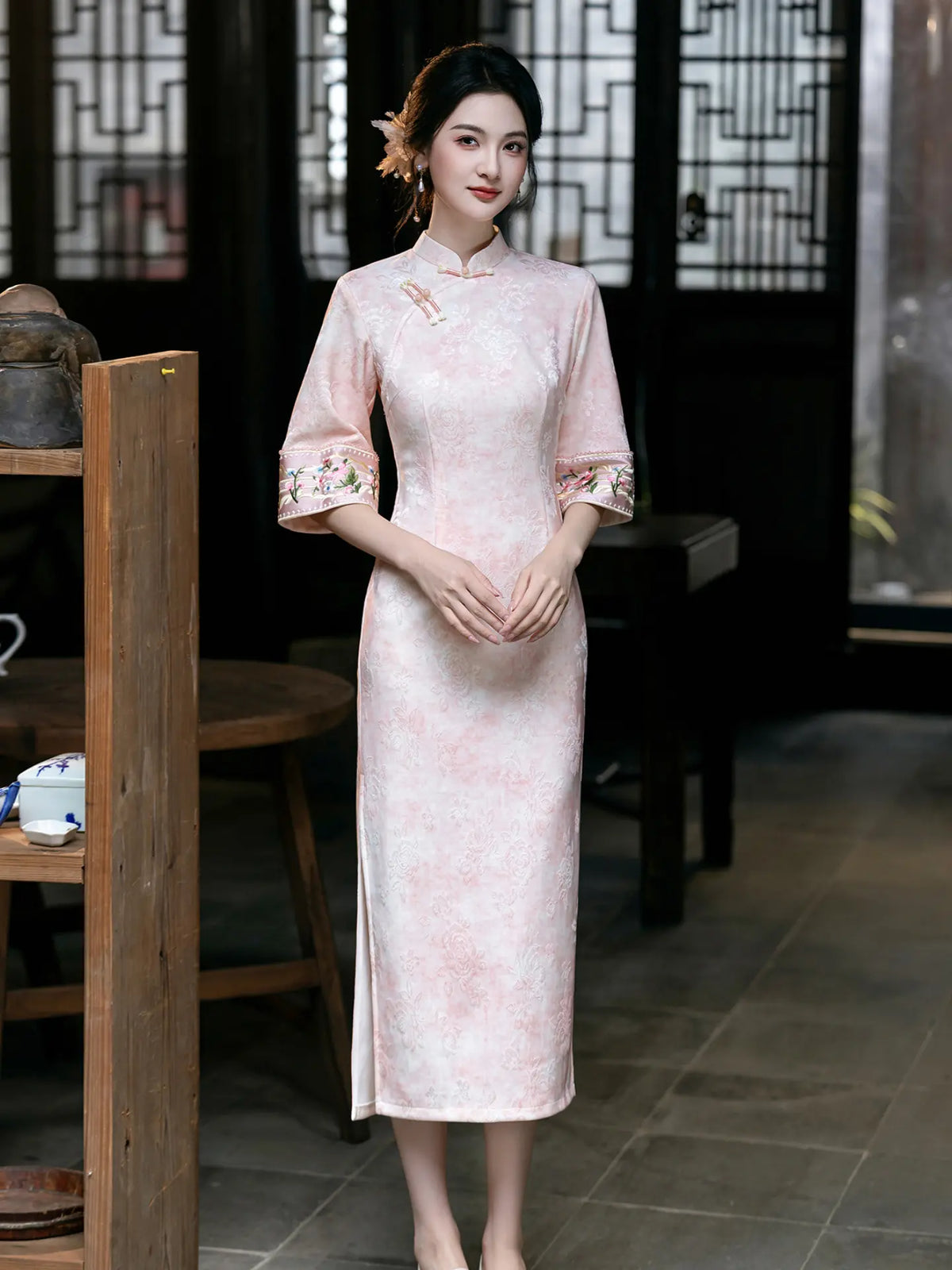 A woman wearing a light pink traditional Chinese cheongsam dress with floral embroidery on the sleeves, standing in a dark, traditional interior setting.