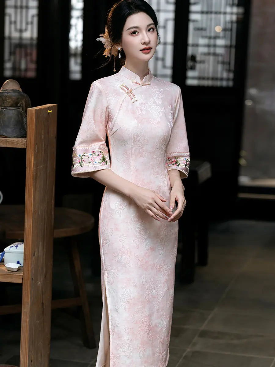 A woman wearing a light pink traditional Chinese cheongsam qipao dress with floral embroidery on the sleeves, standing in a dimly lit indoor setting with wooden furniture.