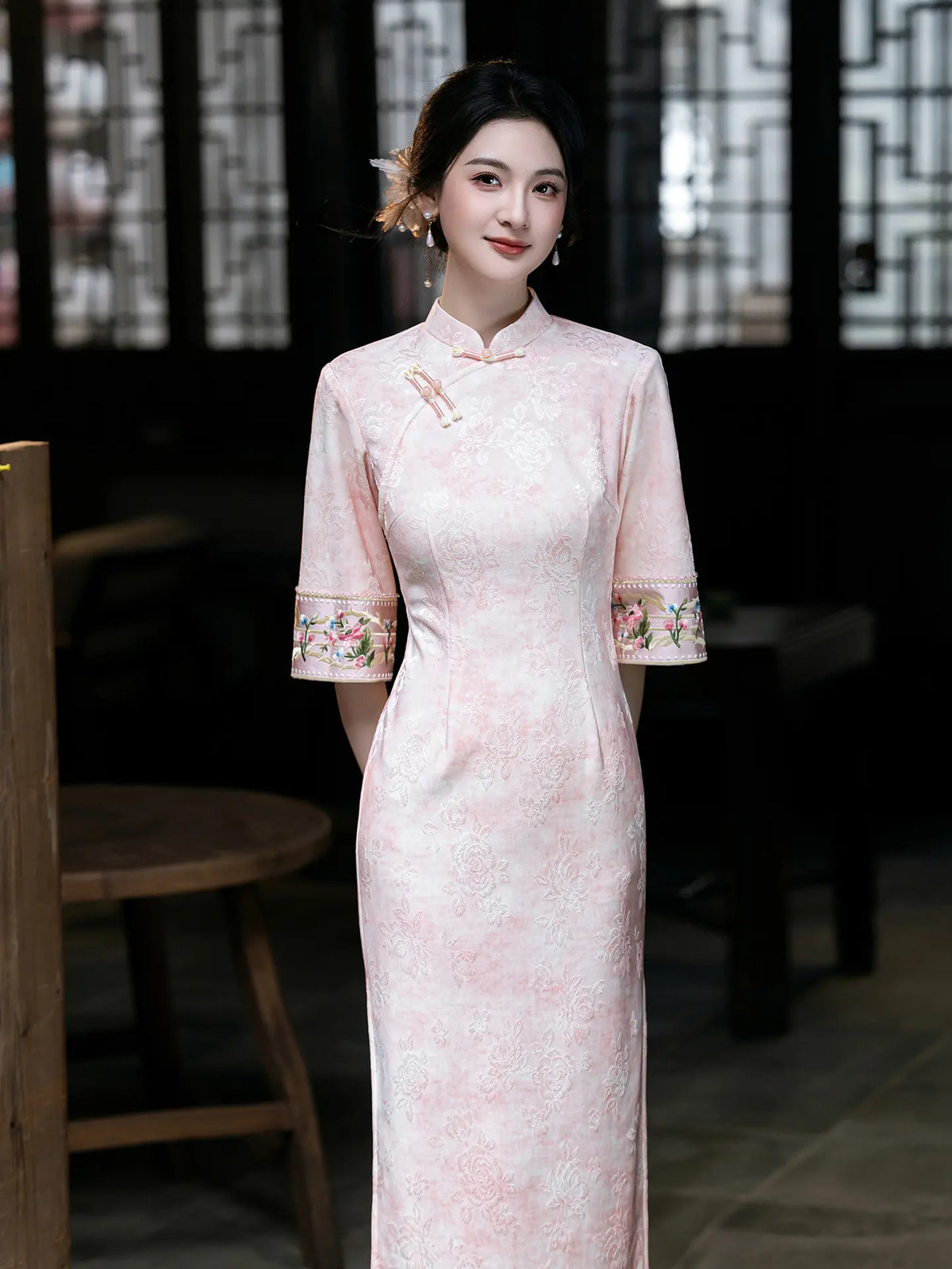 A woman wearing a light pink traditional Chinese cheongsam dress with floral embroidery on the cuffs, standing in a dimly lit traditional room.