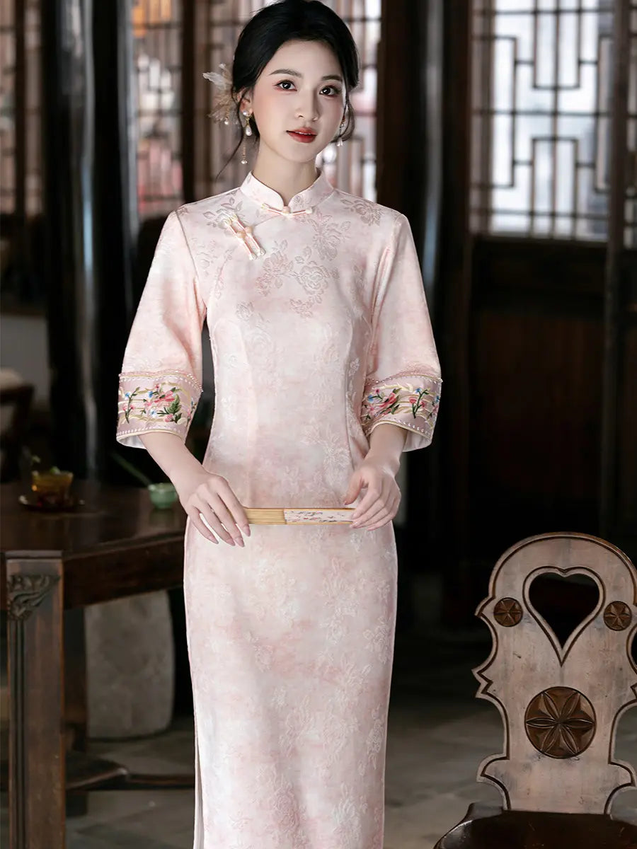 A woman wearing a light pink traditional Chinese cheongsam (qipao) dress with floral embroidery on the sleeves, standing in a traditional Chinese room with wooden lattice windows.