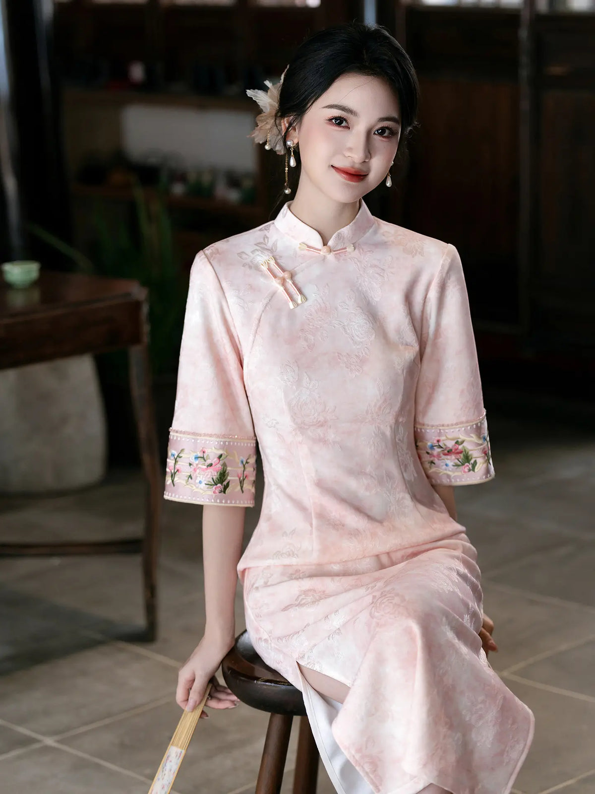 A woman wearing a light pink traditional Chinese cheongsam dress with floral embroidery on the sleeve cuffs, seated on a wooden stool in a dimly lit indoor setting.