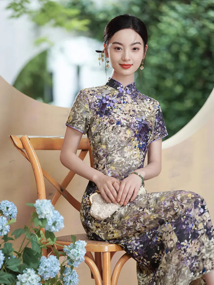 A woman wearing a short-sleeved Botanical Violet & Olive Floral Jacquard Modern A Line Qipao cheongsam dress sits on a wooden chair in an outdoor setting with green foliage and light blue flowers in the foreground.