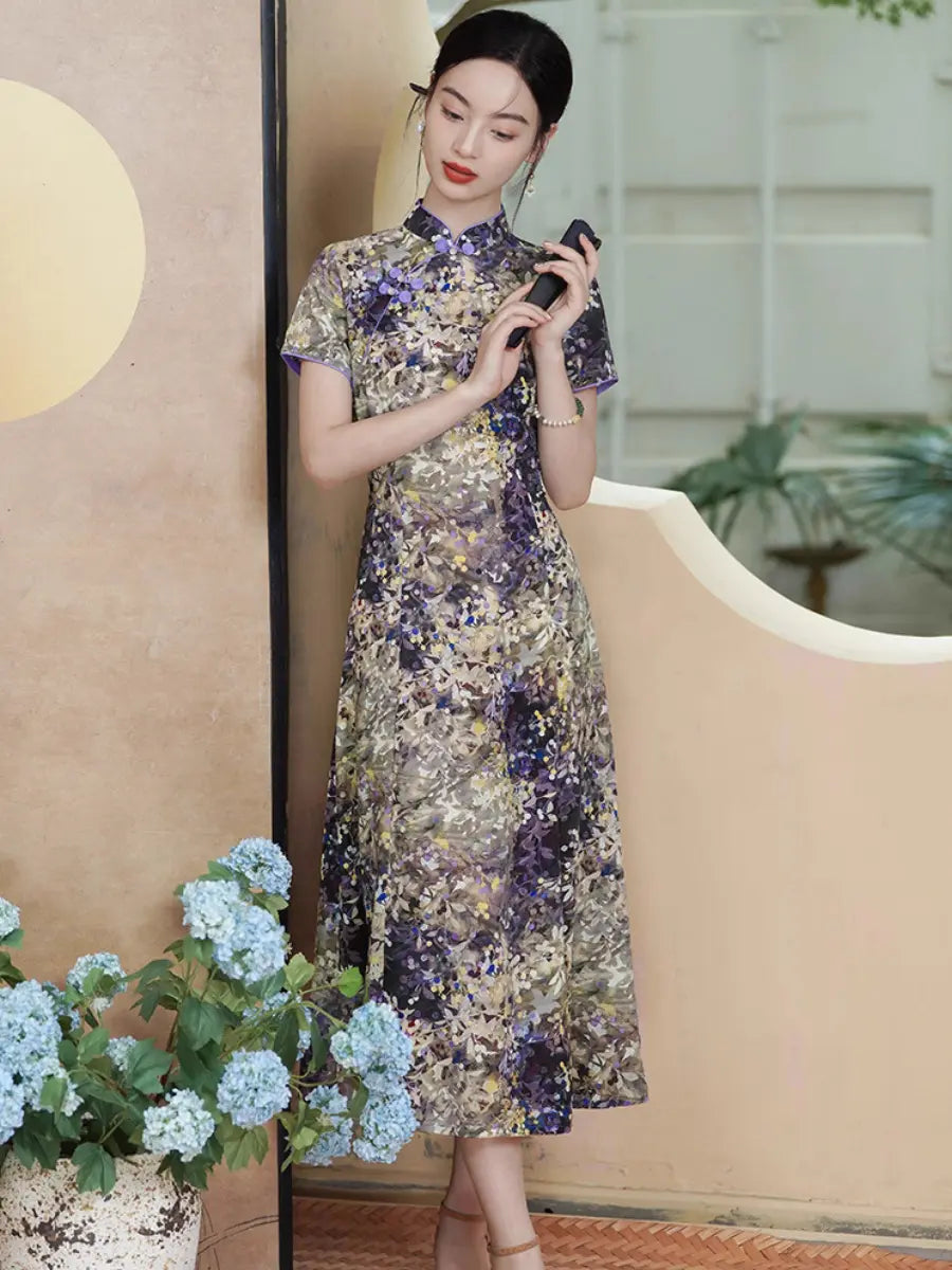 A woman models a short-sleeved  Botanical Violet & Olive Floral Jacquard Modern A Line Qipao cheongsam dress with a dense floral pattern in purple, gold, and beige. She is standing in a styled indoor setting with a beige wall, decorative circular elements, and potted blue hydrangeas in the foreground.