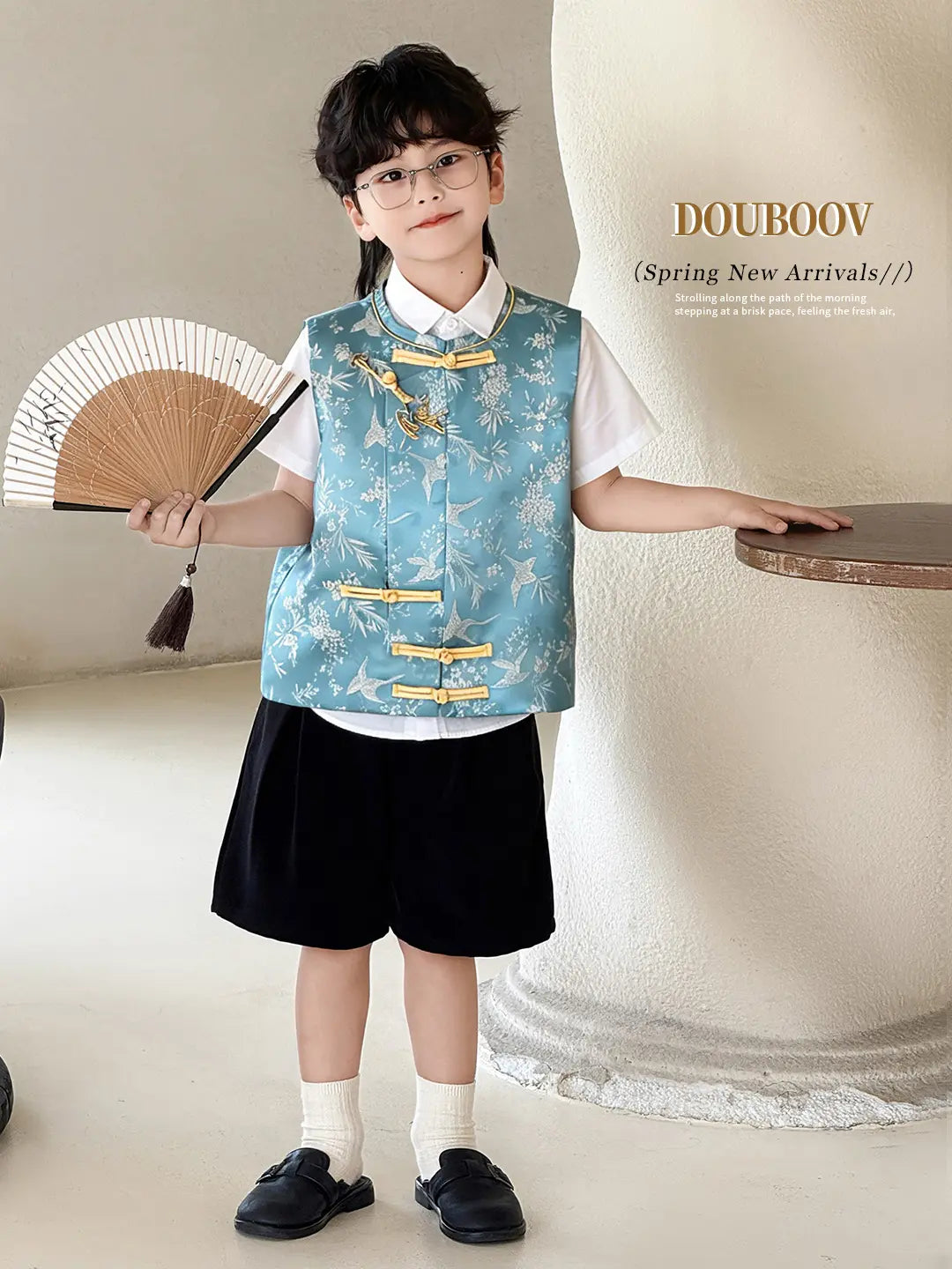 Child wearing a blue floral Boys' Traditional Chinese Style Blue Vest & Shorts Outfit  with gold accents, holding a fan, against a neutral background.