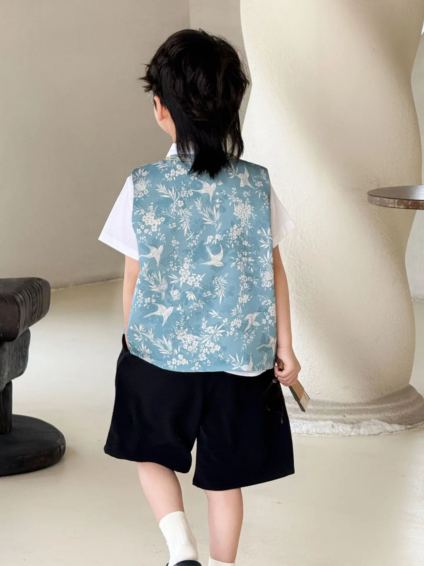 Child wearing a light Boys' Traditional Chinese Style Blue Vest Outfit over a white shirt and black shorts, standing indoors.