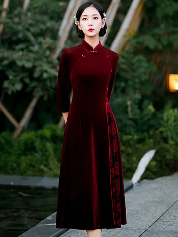 Woman in a Burgundy Red Velvet A line Qipao with Faux Pearl Buttons dress standing outdoors with greenery in the background