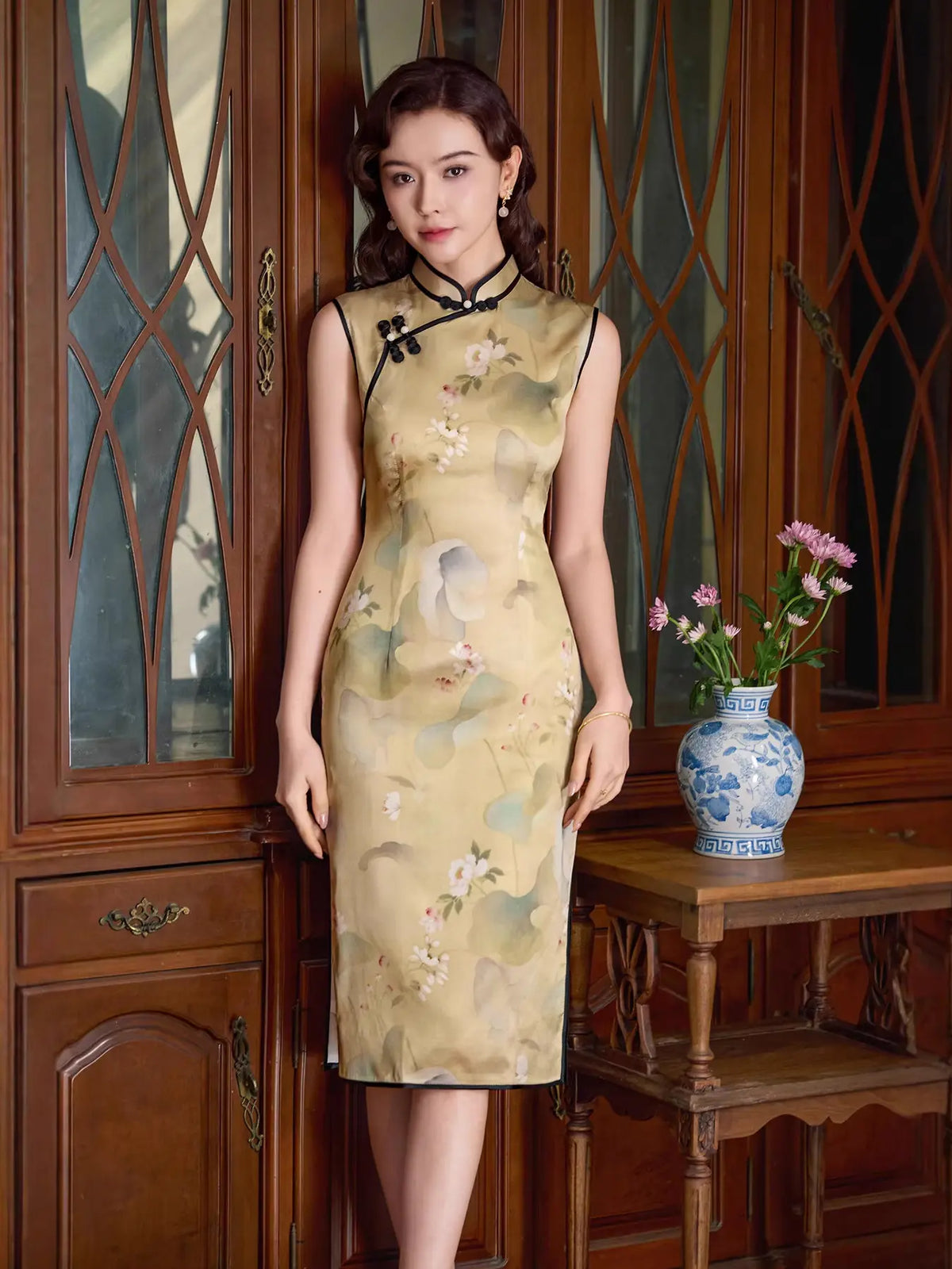 A woman stands indoors wearing a sleeveless beige silk cheongsam dress with a floral and bird print. She is posed in front of a dark wood cabinet, with a small table and blue-and-white vase holding pink flowers to her right.