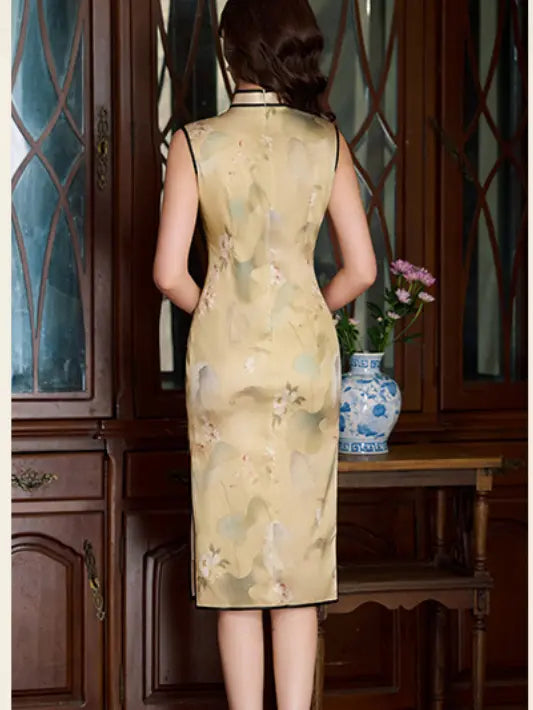 A woman stands with her back to the camera wearing a sleeveless beige silk cheongsam dress with a subtle floral print and black trim, posed in front of a dark wood cabinet and table with a blue-and-white vase.
