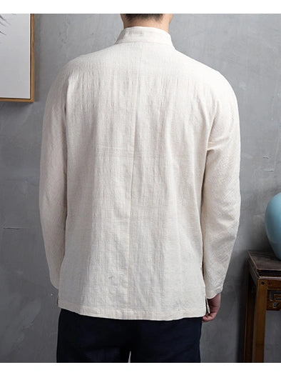 A man viewed from the back wearing a light cream-colored, long-sleeved linen shirt with a mandarin collar, paired with dark pants, set against a textured gray wall.